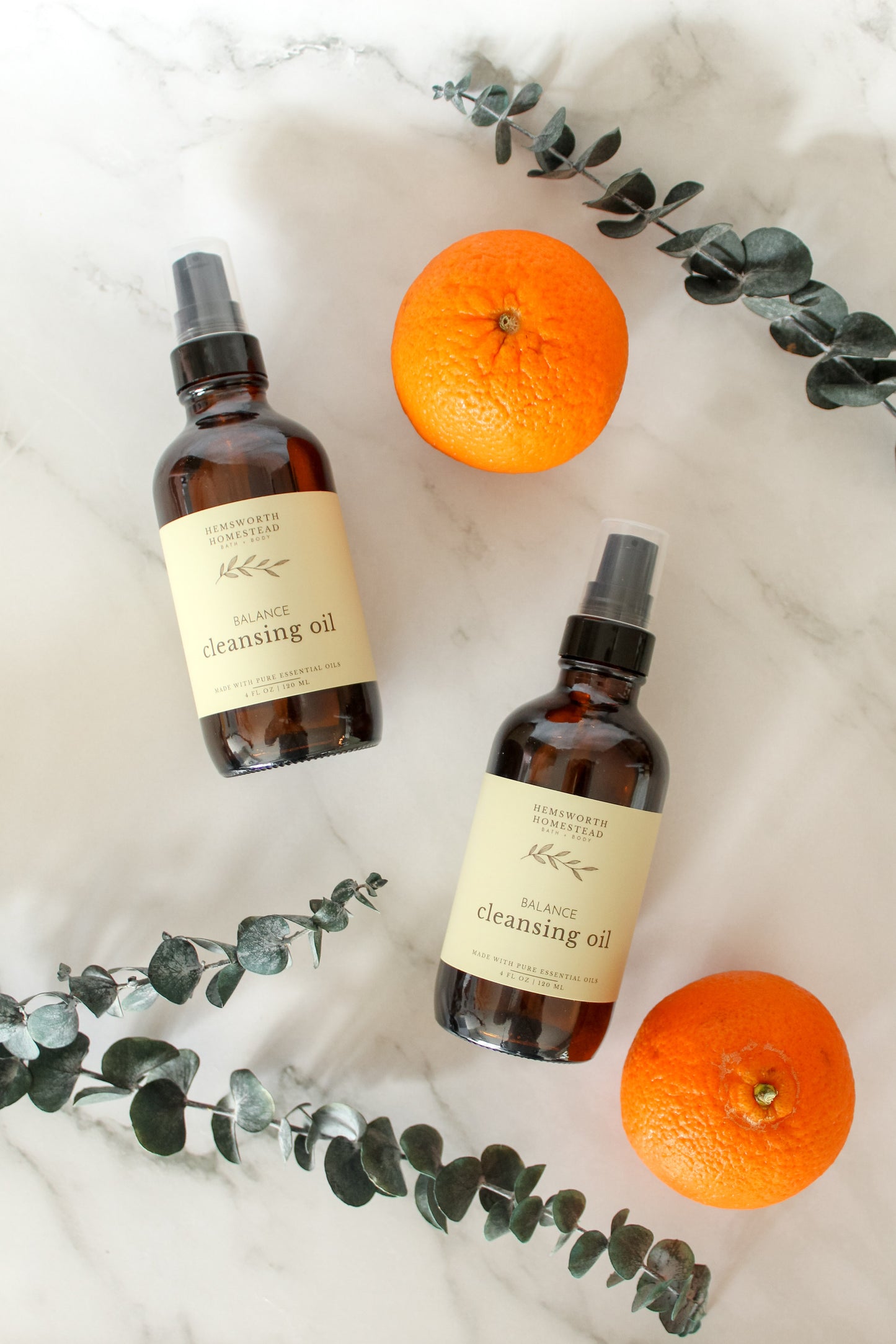 Overhead flat-lay of two Balance Cleansing Oil bottles on marble with fresh oranges and trailing eucalyptus