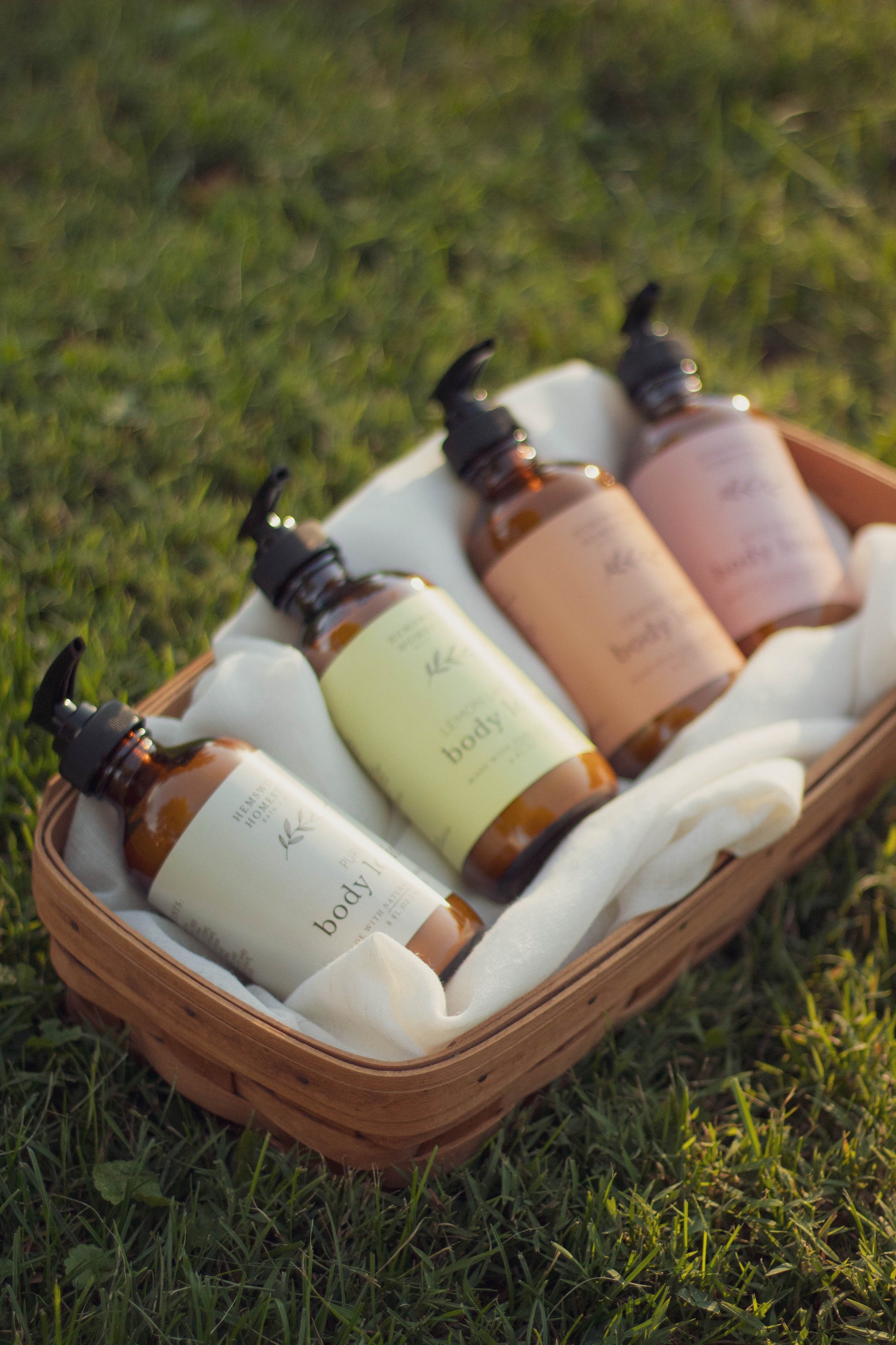 Four bottles of natural body lotion nestled in a picnic basket with a cream cloth, sitting on green grass