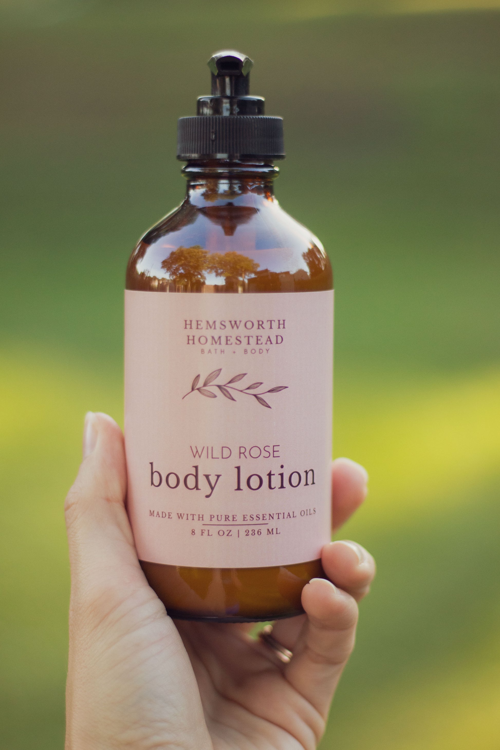 Hand holding a bottle of Wild Rose body lotion with a blush pink label, outdoors in front of green grass