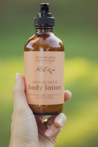 Hand holding a bottle of Orange Grove body lotion with a peach label, outdoors against a green background