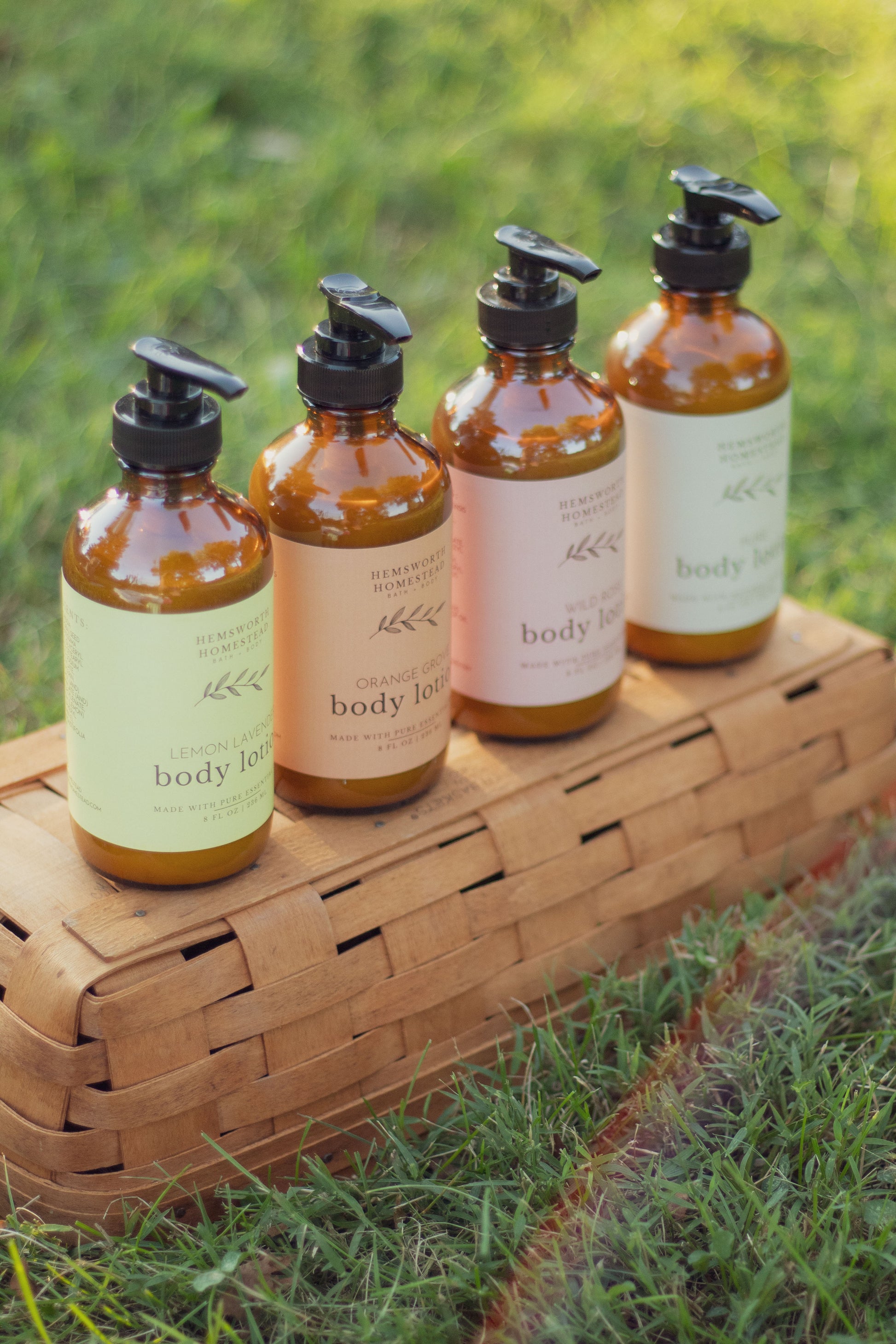 Four amber pump bottles of Hemsworth Homestead body lotion lined up on a woven picnic basket in the grass