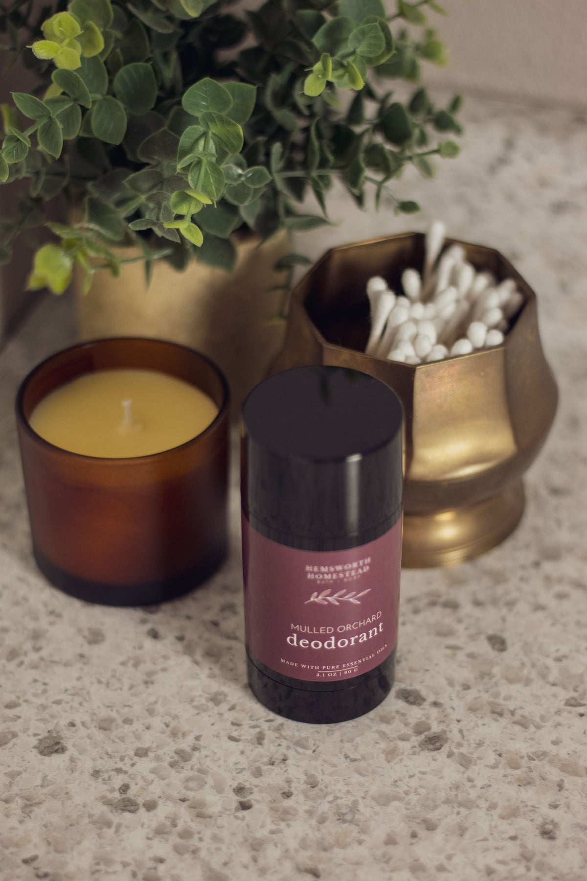 Mulled Orchard deodorant styled with a lit candle, greenery and cotton pads on a marble countertop