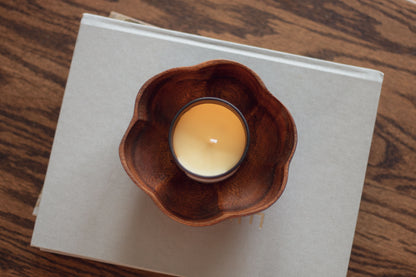 Overhead view of a lit Vanilla Woods candle in a wooden flower-shaped dish on a dark wood table