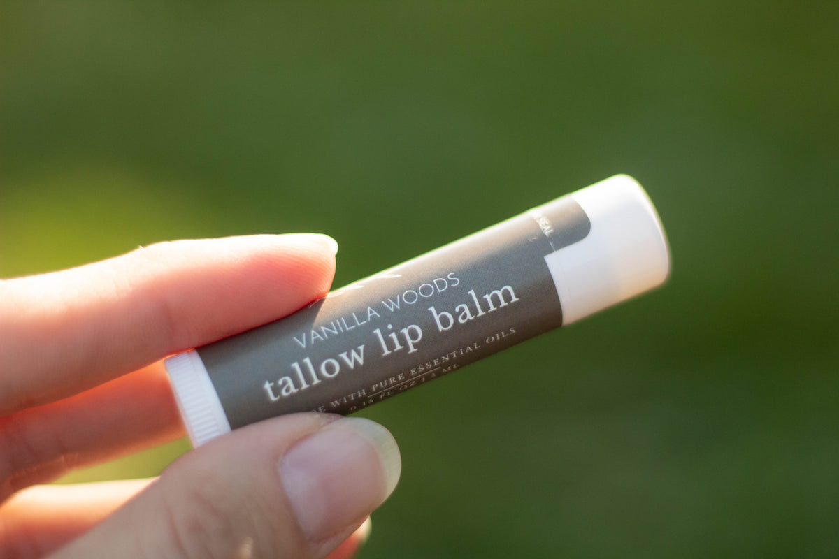 Hand holding a tube of Vanilla Woods tallow lip balm outdoors with a blurred green background