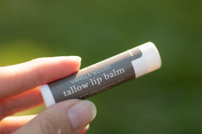 Hand holding a tube of Vanilla Woods tallow lip balm outdoors with a blurred green background