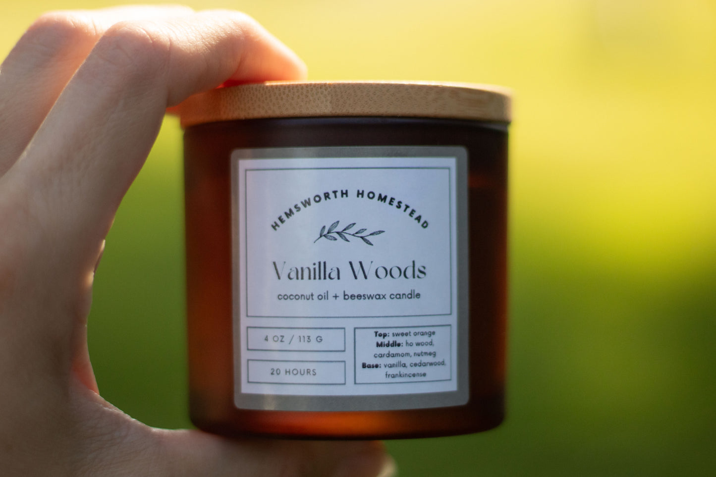 Hand holding an amber glass Vanilla Woods candle jar outdoors with a blurred green background