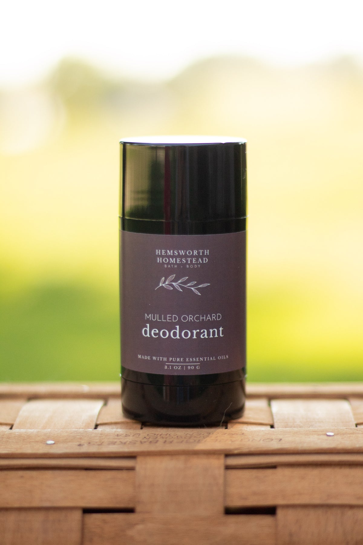 Mulled Orchard deodorant stick standing on a woven basket surface with a blurred green background